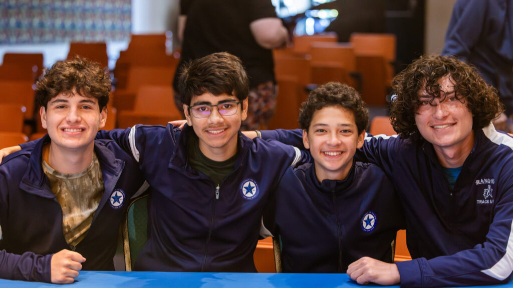 Four young boys are seated at a table, smiling and posing for the camera. They are wearing matching navy blue jackets with a star emblem. The background features rows of orange chairs and a blurred figure in the distance.