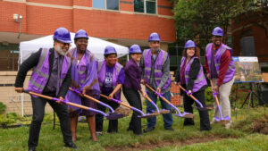 A group of six people in purple hard hats and vests are participating in a ceremonial groundbreaking event. They are holding shovels and posing together on a grassy area, with a tent and a building in the background.