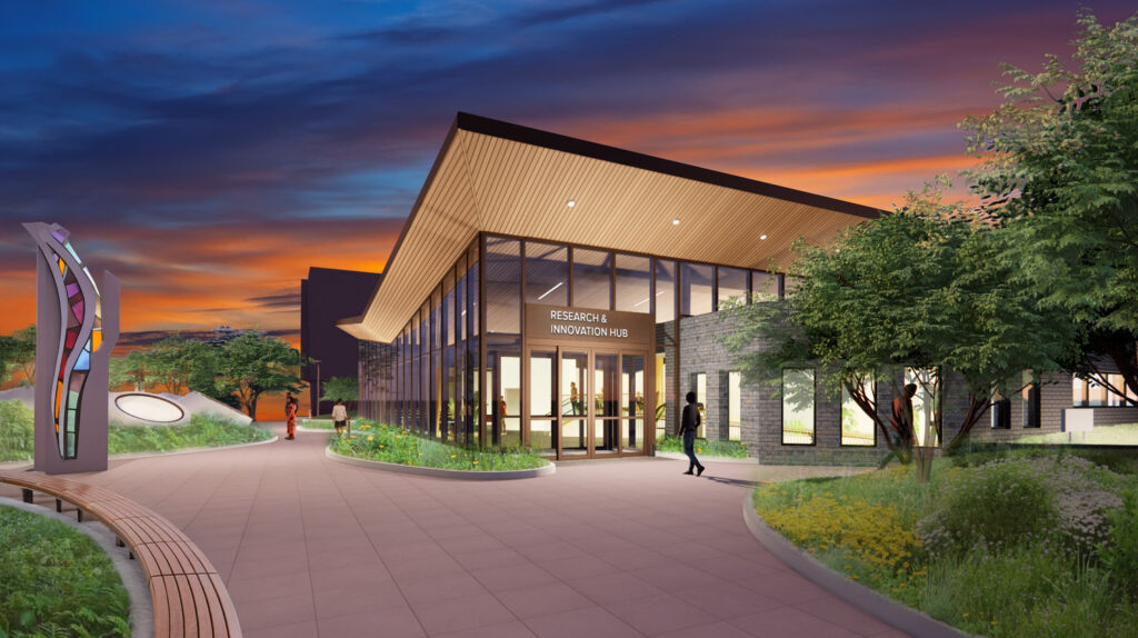 A modern building with large glass windows and a sloping roof is set against a vibrant sunset sky. The pathway leading to the entrance is lined with greenery, and a person walks toward the door. In the foreground, a colorful sculpture is visible, adding to the inviting atmosphere of the scene.