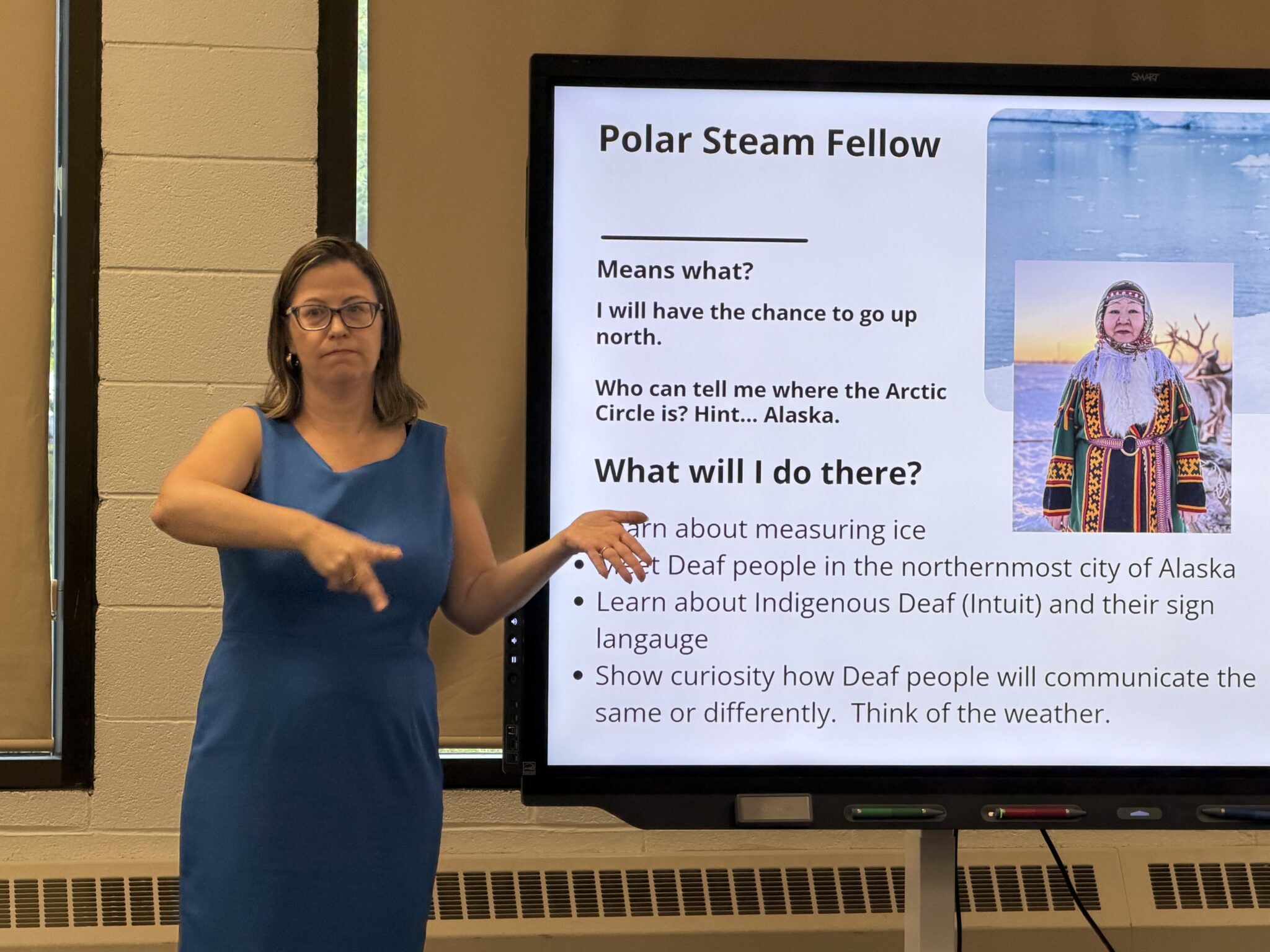 Woman in a blue dress stands next to a screen that shows the image of an Inuit woman. The text on the screen reads, "Polar Steam Fellow. Means what? I will have the chance to go up north. Who can tell me where the Arctic Circle is? Hint...Alaska. What will I do there? Learn about measuring the ice. Meet Deaf people in the northernmost city in Alaska. Learn about Indigenous Deaf (Inuit) and their sign language. Show curiosity how Deaf people will communicate the same or differently. Think of the weather."
