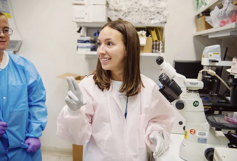 Woman in lab coat and gloves stands next to a microscope and is signing something to a person off to the left.