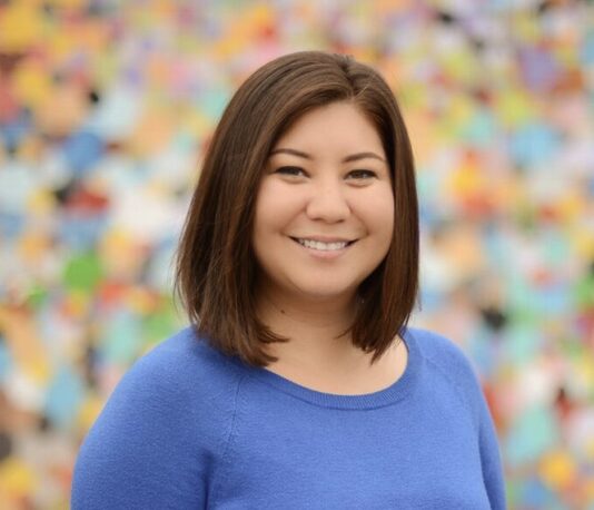 Woman with dark, shoulder-length hair wears a blue top and stands in front of a multi-colored backdrop