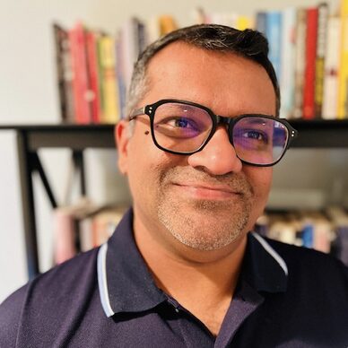 Man with glasses wears a navy blue polo shirt and stands in front of a bookcase