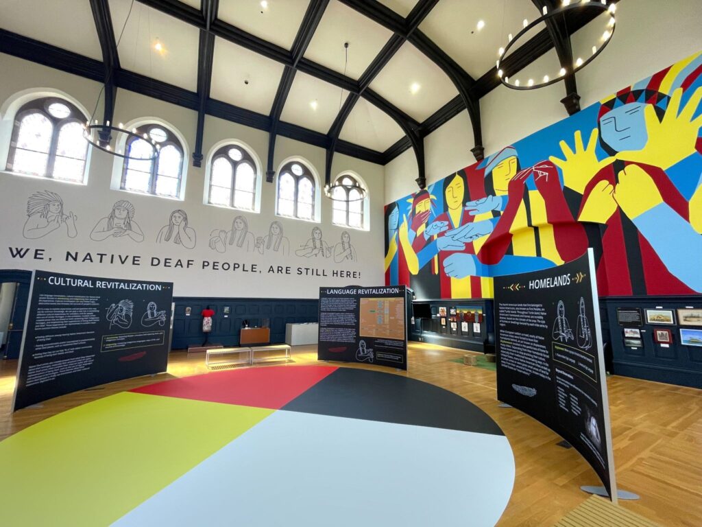 An indoor exhibition space featuring large colorful murals on the walls, including a prominent mural with figures in red, yellow, and blue. The floor is divided into sections of different colors—black, red, yellow, and blue. Several display panels with text are arranged around the room, and there are large windows letting in natural light. A banner with the text "WE, NATIVE DEAF PEOPLE, ARE STILL HERE" is visible on the wall.