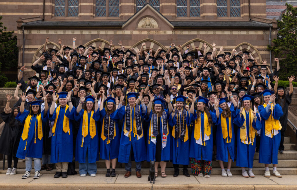 A large group of graduates wearing blue caps and gowns stands outside a building, smiling and raising their hands in celebration. Some graduates are wearing additional honors cords and stoles. The background features a brick building with large windows and greenery.