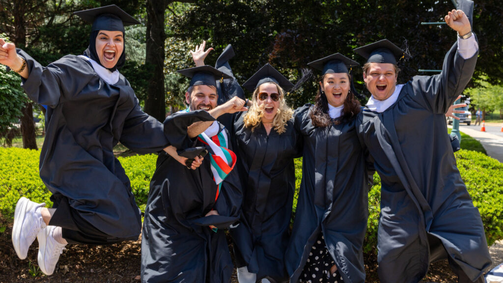 A group of five graduates in caps and gowns joyfully posing outdoors, with one person jumping in the air and the others smiling and celebrating. They stand in front of lush greenery, with some wearing stoles or accessories that signify their achievements.